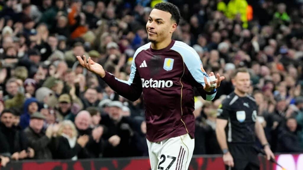 Footballer Morgan Rogers playing for Aston Villa Football Club in a maroon and blue shirt with white shorts bearing his number 27 number in black lettering. Rogers is in front of a large crowd with his arms outstretched