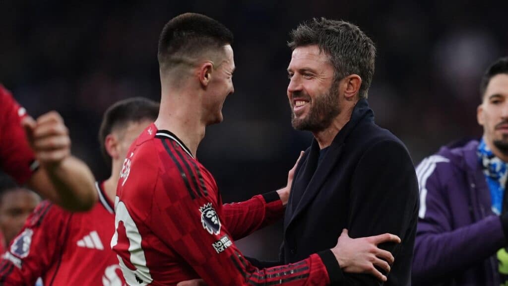 Manchester United Football Club manager Michael Carrick congratulates one of his players while facing to his left, smiling and wearing a black top. Carrick has dark hair and a moustache and beard. His player is wearing a red, black and white Man United shirt, with other players visible on a football pitch