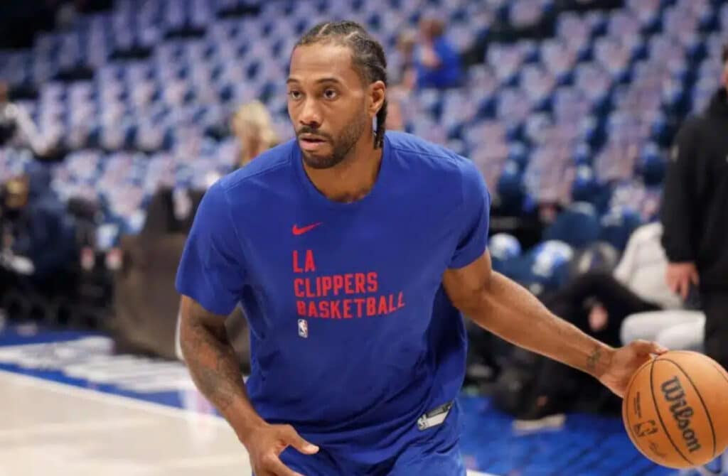 NBA basketball player Kawhi Leonard holding an orange ball with the word Wilson in black on it on a court in front of empty seats. Leonard is looking to his right and wearing a blue t-shirt with the words Los Angeles Clippers on it in red. People are blurred but visible in the background