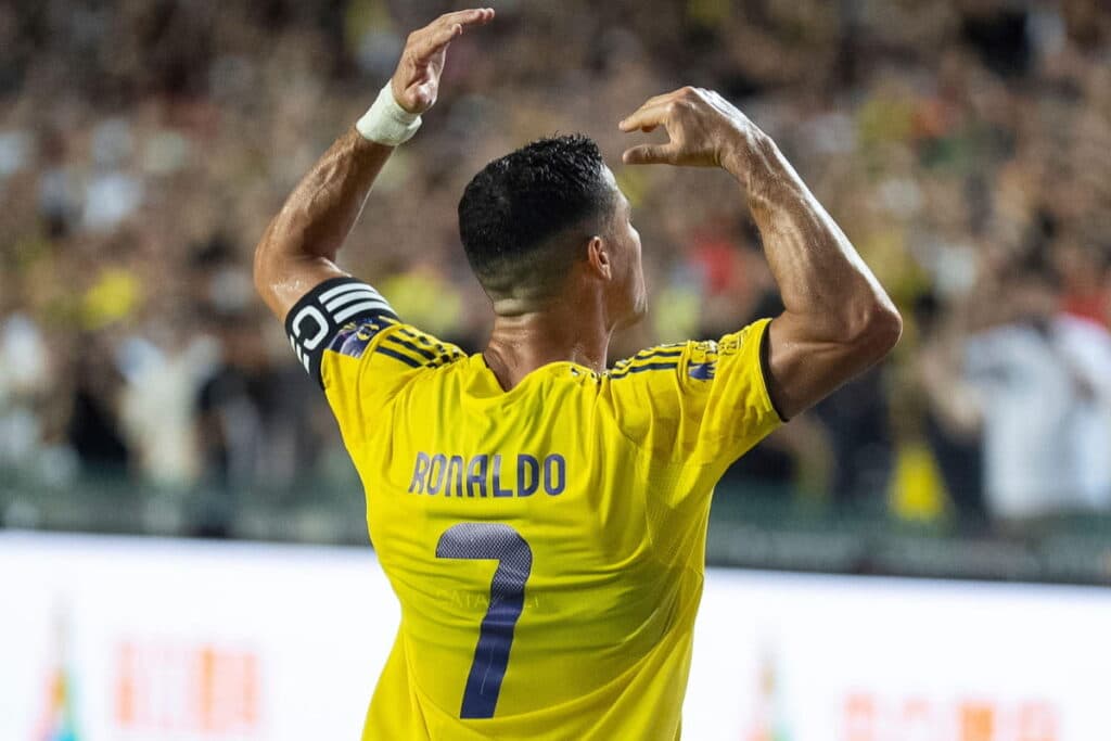 Footballer Cristiano Ronaldo playing football for Saudi Pro League club Al Nassr. Ronaldo is holding his arms in the air in a yellow shirt with a captain's armband and his name and number 7 number in blue, as well as blue trim. A blurry crowd is visible