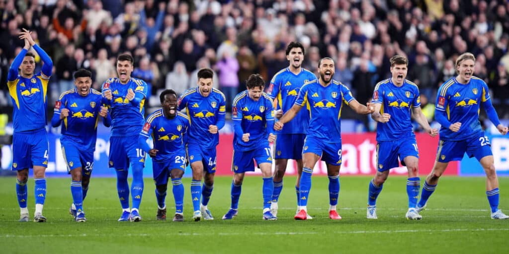 Leeds United Football Club footballers Lucas Perri, Jayden Bogle, Joe Rodon, Jaka Bijol, Pascal Struijk, Ethan Ampadu, Ao Tanaka, Brenden Aaronson, Sebastiaan Bornauw, Ilia Gruev and Dominic Calvert-Lewin standing in a line celebrating after winning a penalty shootout in the FA Cup. The players are gesturing and embracing each other in a predominantly blue kit with yellow trim and different coloured boots. A packed crowed is blurrily visible in the background