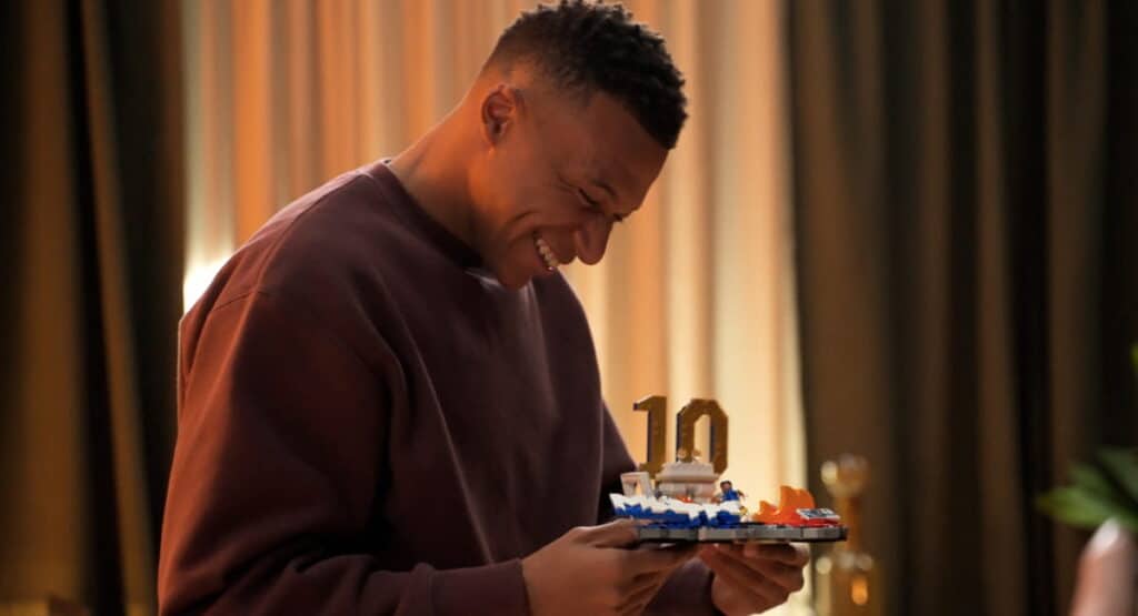 Footballer Kylian Mbappe grinning while holding up a lego toy carrying the number 10 in a darkened room. Mbappe is wearing a dark-coloured top
