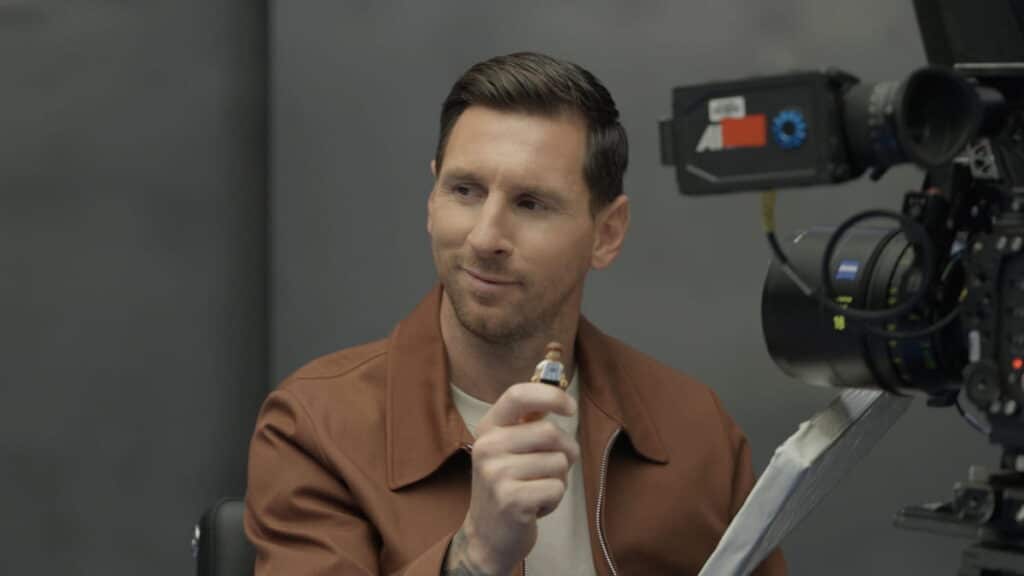 Footballer Lionel Messi sitting in a room while being filmed. A camera is visible in the foreground and messi is wearing a brown jacket and white t-shirt while holding up a hand
