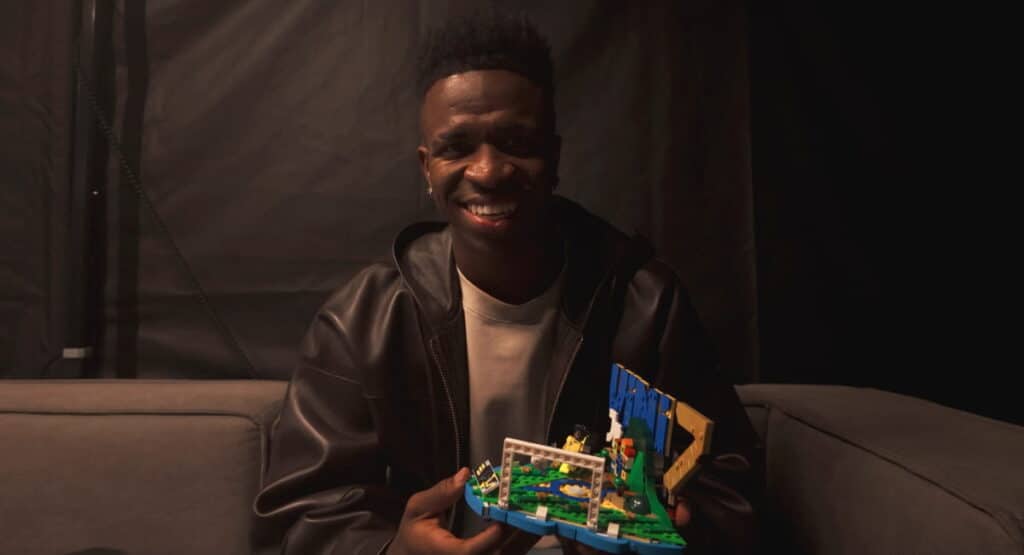 Footballer Vinicius Junior grinning while holding up a lego toy in a darkened room. Vinicius Junior is wearing a dark-coloured jacket